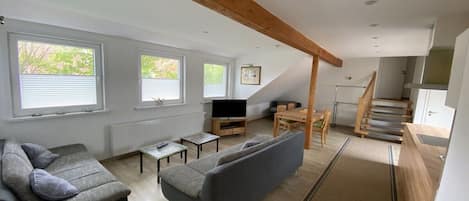 Sala de estar moderna com sofá, televisão e área de jantar, destacando teto inclinado e vigas de madeira.