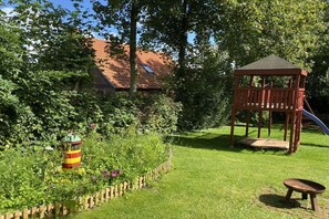 A garden with a wooden play structure, a colorful fire hydrant, and a wheelbarrow. - Holiday apartment with 2 bedrooms (Pilsum)
