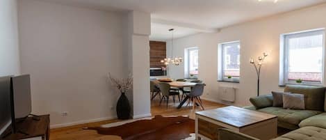 Un salon moderne avec un canapé, une table basse et un téléviseur. On aperçoit à l’arrière-plan un coin repas avec des chaises et une table.