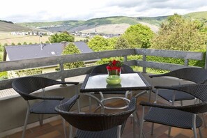 Een balkon met rieten stoelen en een tafel, met uitzicht op een dorp en glooiende groene heuvels.