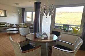 Een moderne woonkamer met een ronde glazen tafel, omringd door rieten stoelen, een decoratieve metalen kroonluchter en uitzicht op een veld door grote ramen.
