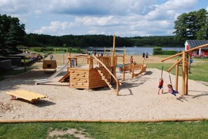 A playground with wooden structures, including slides and climbing areas, near a body of water. - Holiday island Tietzowsee - Finnish house (Zechlinerhütte)