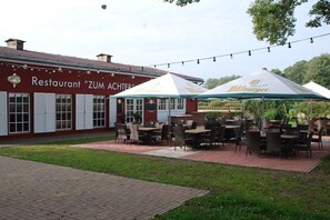 An outdoor seating area with tables and chairs, a red building with a sign, and string lights. - Holiday island Tietzowsee - Finnish house (Zechlinerhütte)