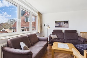 A living room with a brown sofa, a wooden coffee table, and a window with a view of a house. - Holiday apartment Uhlenkamp 4 (Langeoog)