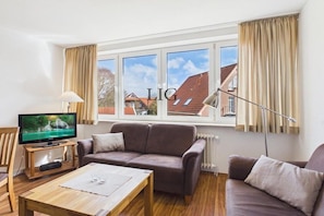 A living room with a television, sofa, and a window with curtains. - Holiday apartment Uhlenkamp 4 (Langeoog)