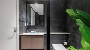 Un baño moderno con un espejo grande, un lavabo con artículos de aseo, un inodoro y un cubo de basura.