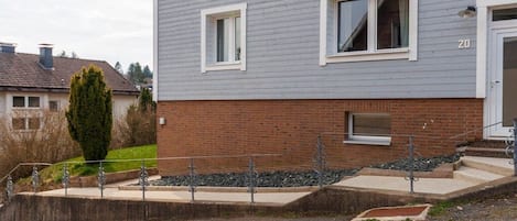 Ein zweistöckiges Wohnhaus mit grauem Außenputz, weißen Fensterrahmen und einem Ziegelunterbau.