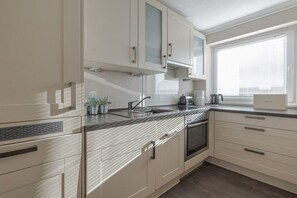 A modern kitchen with white cabinetry, a stainless steel range hood, and a window with blinds. - Margarethenstraße Holiday Apartment 10 (Sylt\/Westerland)