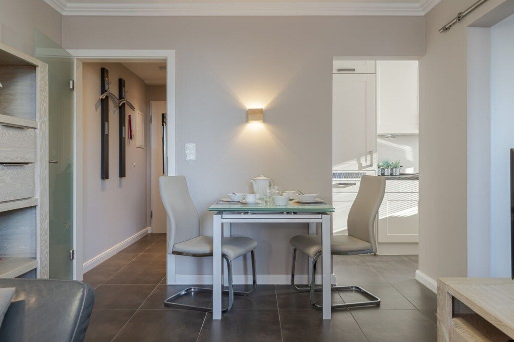 A modern dining area with a glass table, two chairs, and a wall-mounted light fixture.