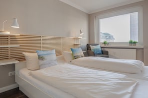 A hotel room with two beds, a window with blinds, a radiator, and a view of a building outside. - Margarethenstraße Holiday Apartment 10 (Sylt\/Westerland)