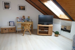 A room with a sloped ceiling, a television on a wooden stand, a wicker chair, a small table with items, and a potted plant. - Holiday apartment with 1 bedroom (Wunstorf\/ Steinhude)