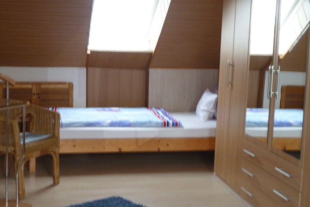 Une chambre avec un plafond en pente, un lit avec un matelas blanc et une couverture à rayures, une armoire en bois et une chaise en osier.