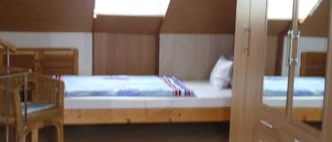 A bedroom with a sloped ceiling, a bed with a white mattress and a striped blanket, a wooden wardrobe, and a wicker chair.
