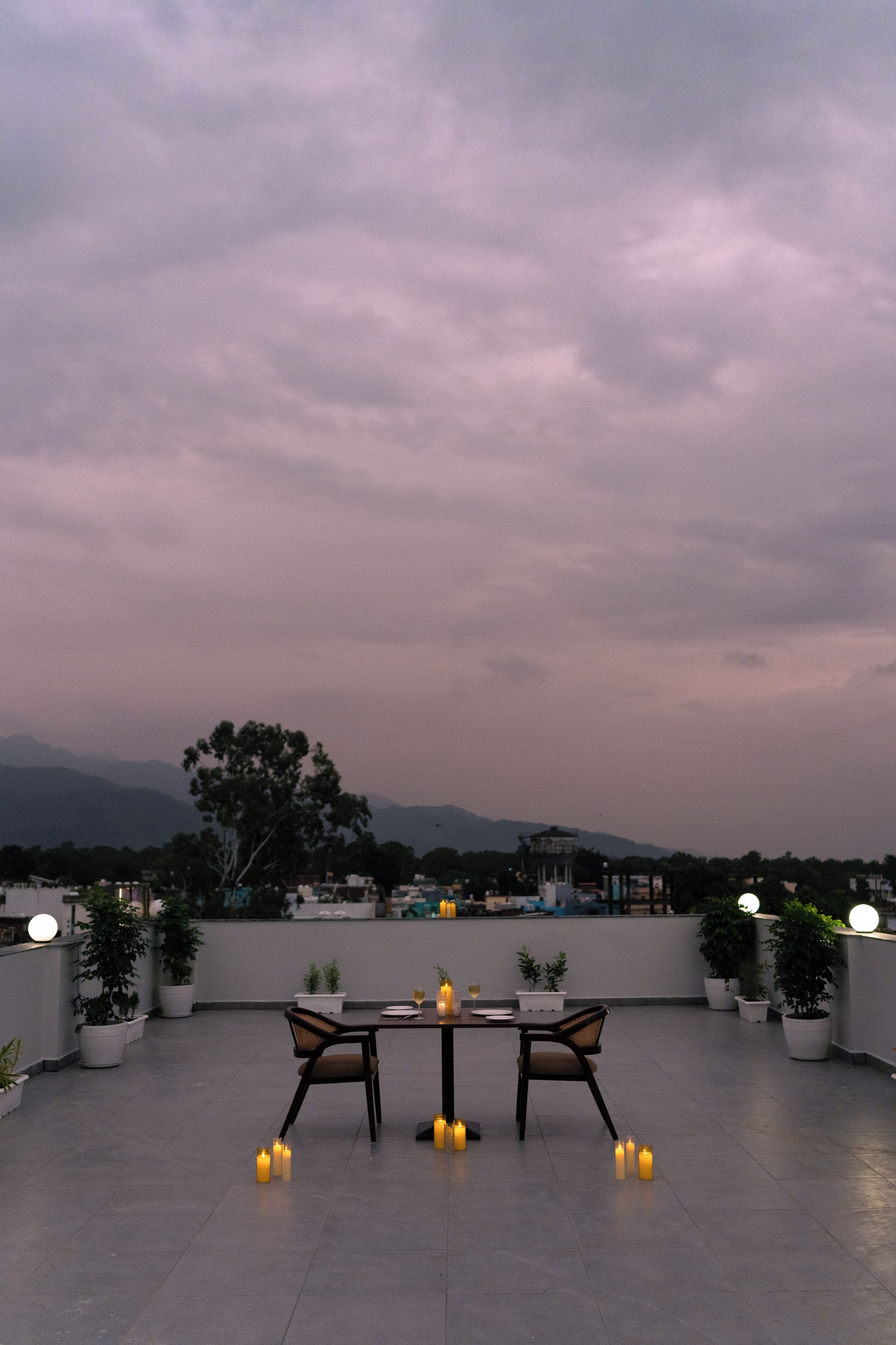 An outdoor seating area with a table and chairs, surrounded by potted plants, under a twilight sky.