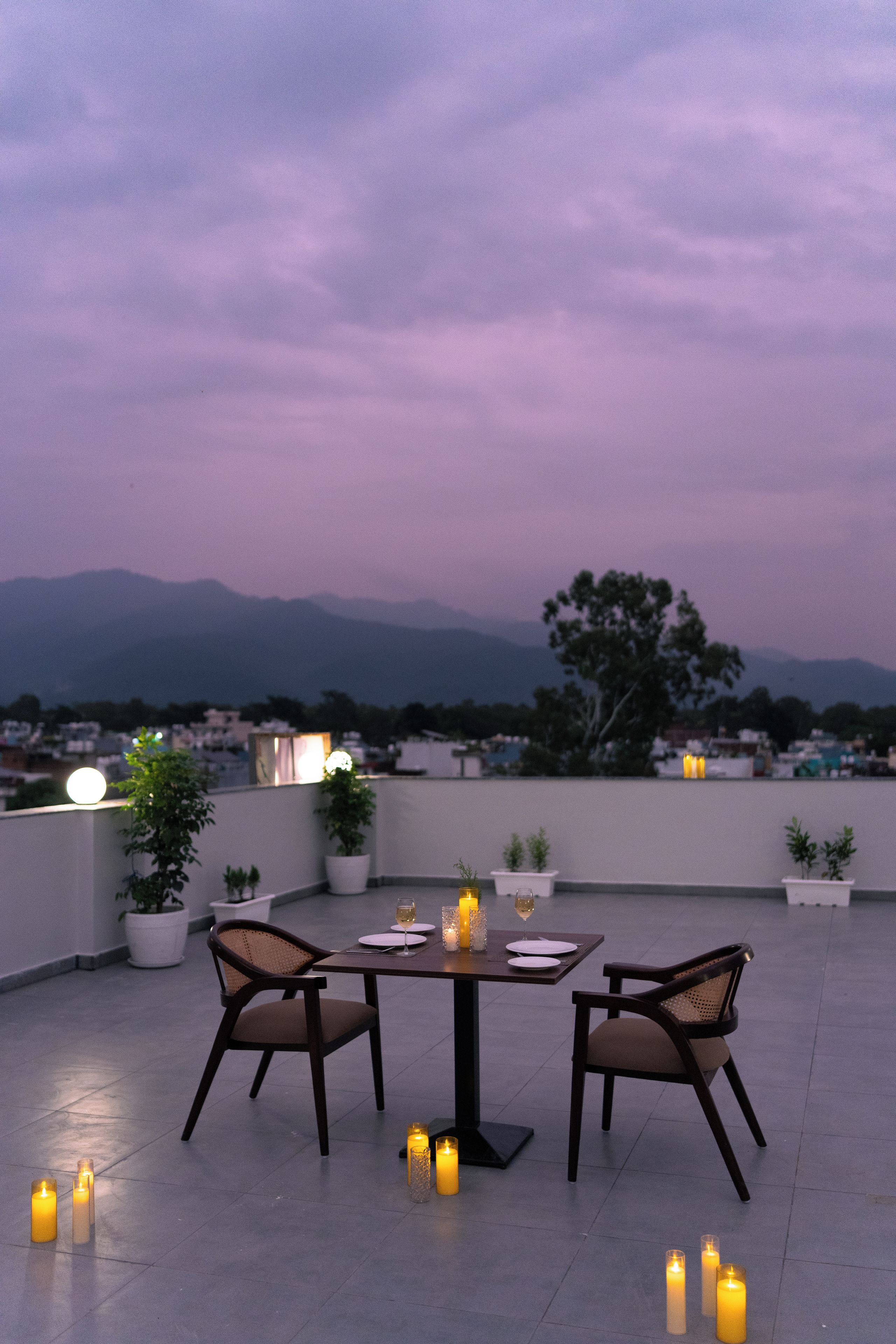 Una terraza en la azotea con una mesa dispuesta para dos, rodeada de velas, plantas en macetas y vistas a un paisaje urbano con montañas al fondo.