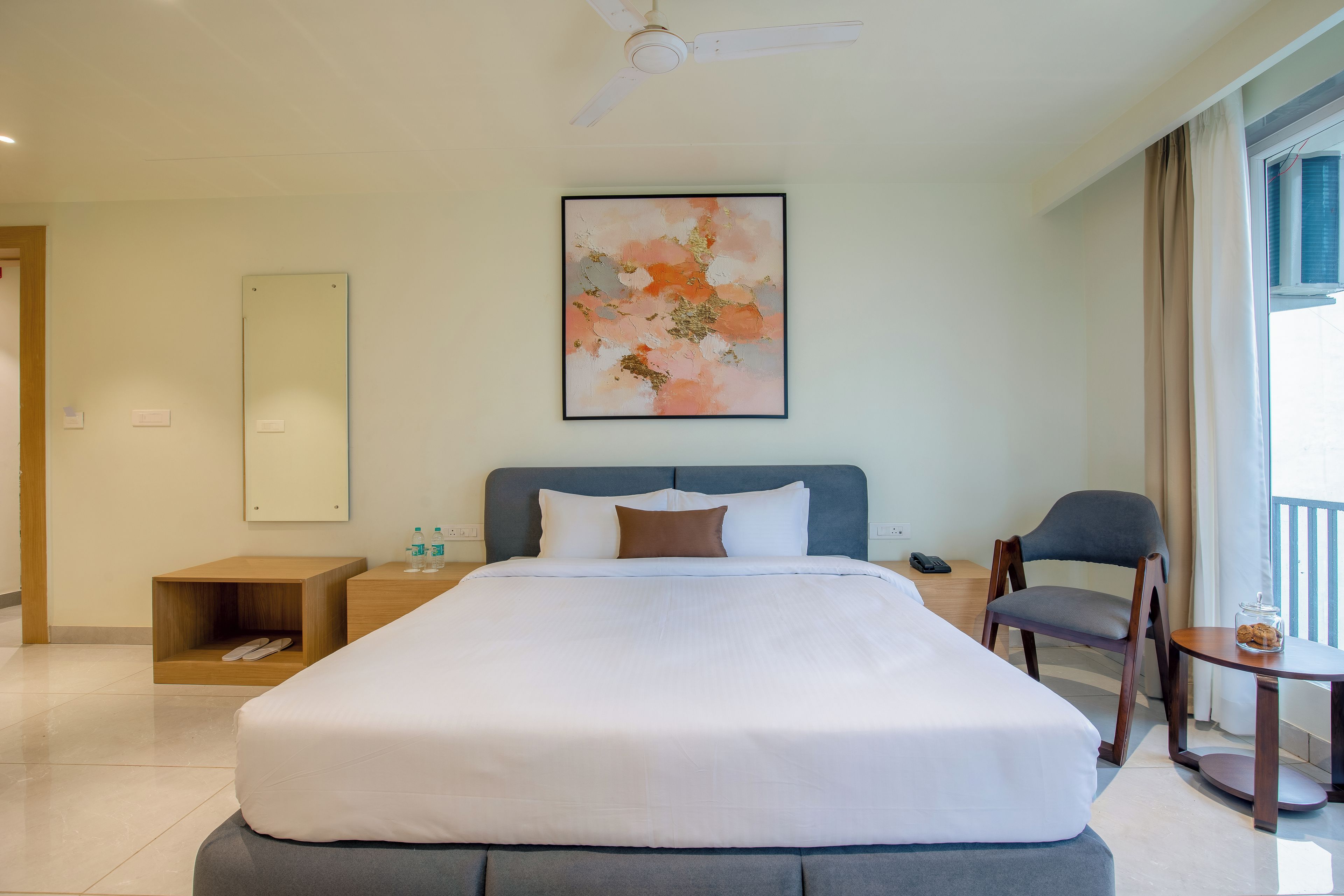 A hotel room with a bed, a chair, a small table, a nightstand, a wall-mounted artwork, and a ceiling fan.