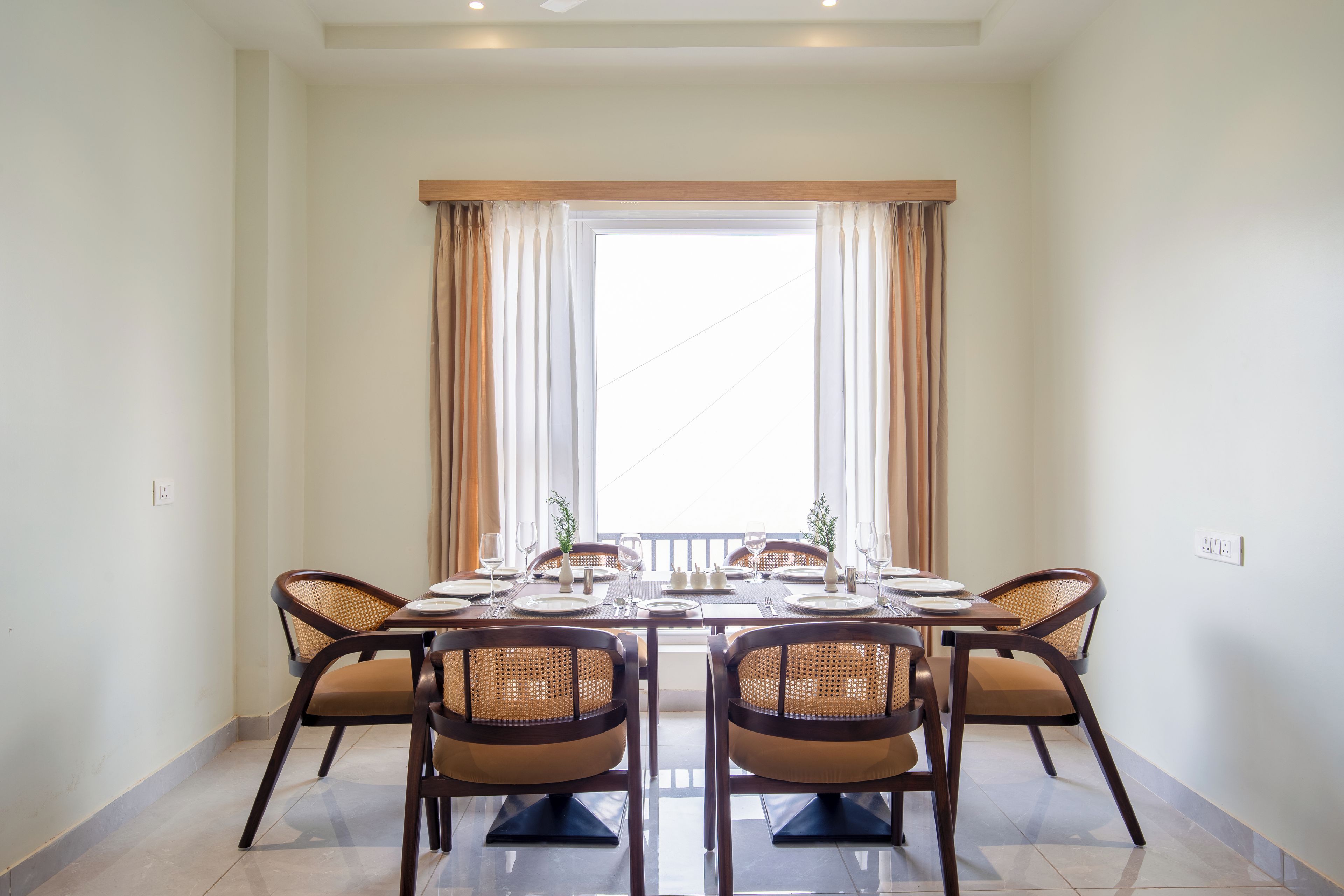 Un comedor con una mesa dispuesta para seis personas, rodeada de sillas de mimbre, un ventanal con cortinas y vistas al exterior.