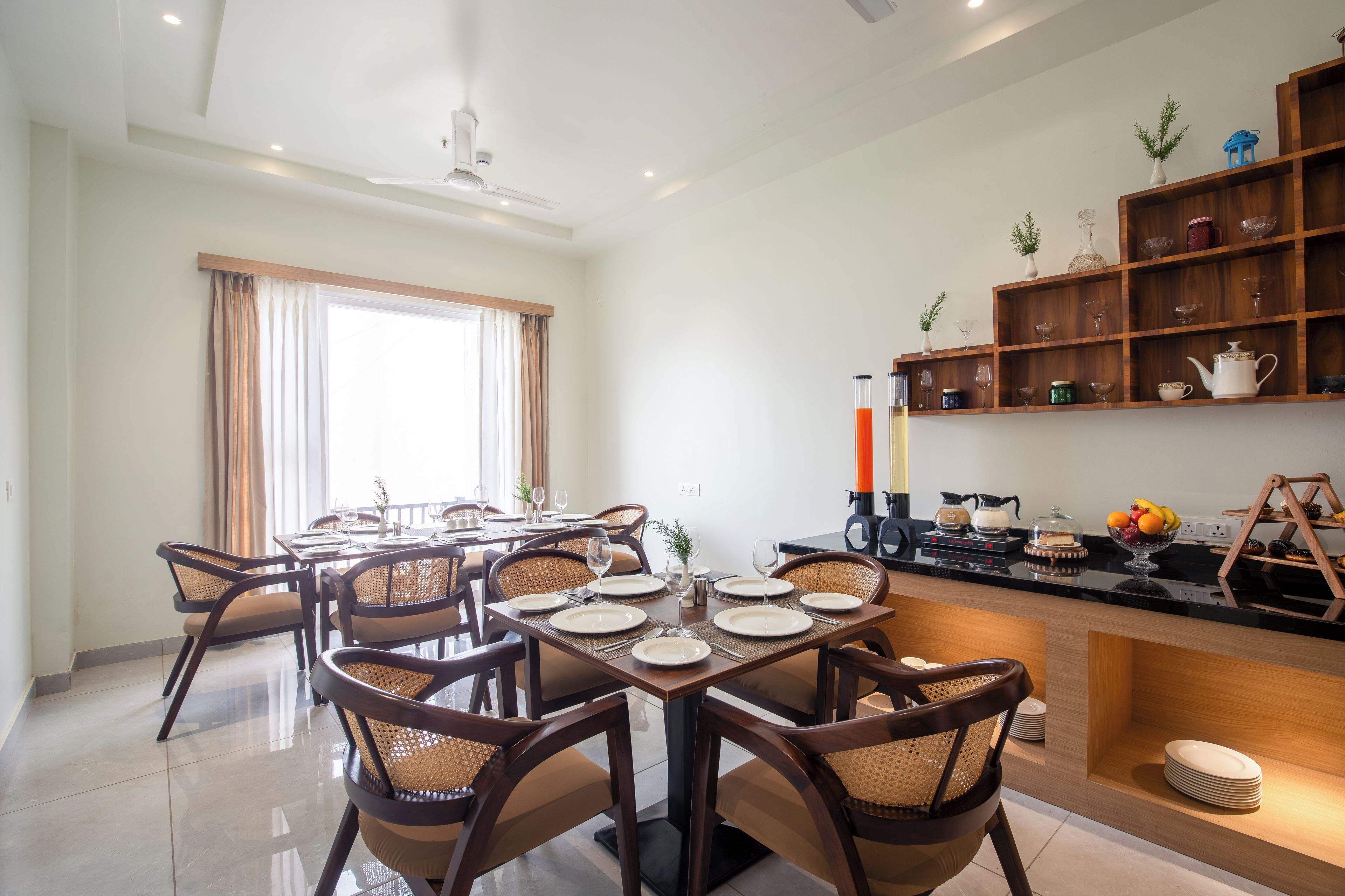 Comedor con una mesa preparada para cuatro personas, rodeado de sillas de mimbre, y una zona de cocina con cafetera y juego de té.
