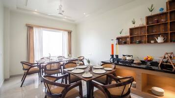 Comedor con una mesa preparada para cuatro personas, rodeado de sillas de mimbre, y una zona de cocina con cafetera y juego de té.