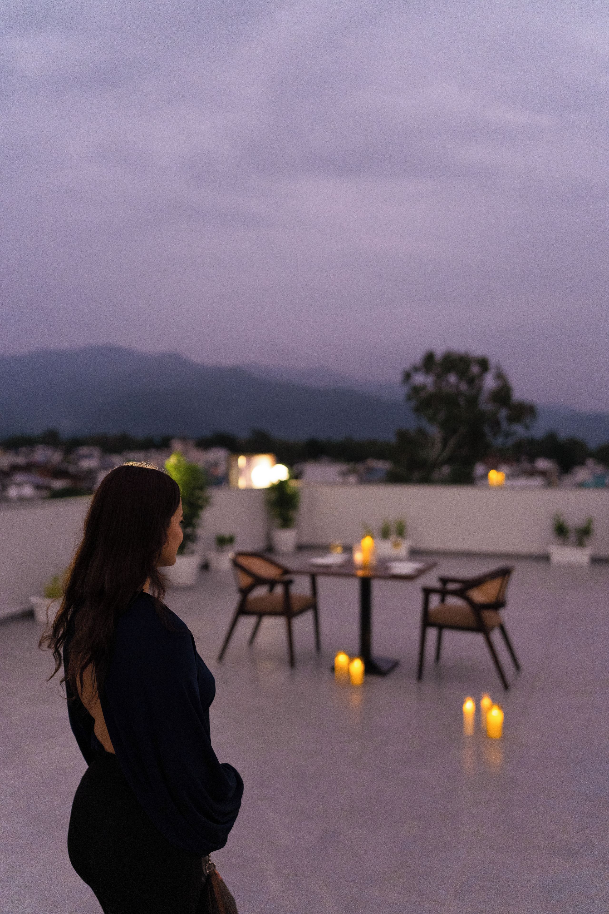 Eine Frau steht auf einer Dachterrassen mit Tisch und Stühlen und blickt auf eine Stadtlandschaft und Berge.