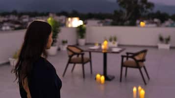 Una mujer de pie en una terraza en la azotea, con una mesa y sillas, contemplando un paisaje urbano y montañas.