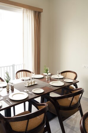 A dining area with a table set for six, surrounded by wicker chairs, a vase with greenery, and a window with curtains. - The Halt Stays (Haldwani)