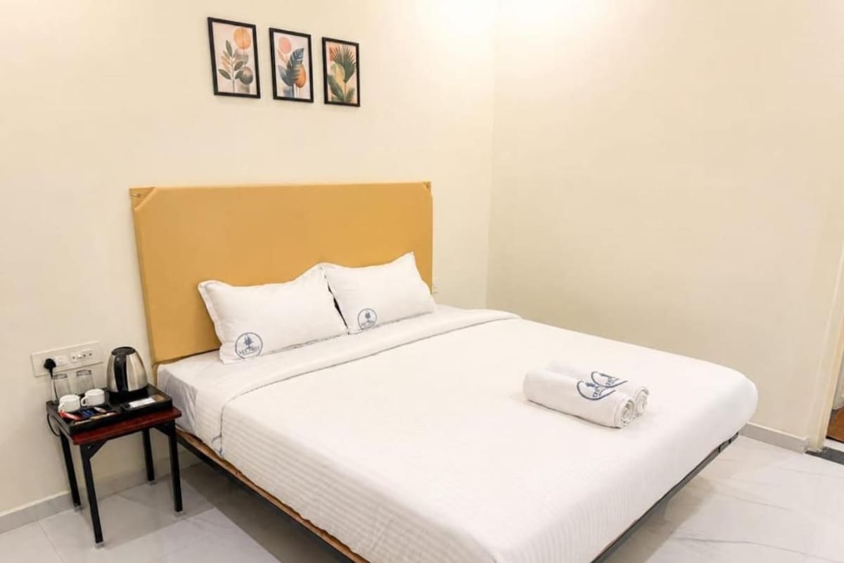 Habitación de hotel con cama, mesita de noche con hervidor y enchufe en la pared.