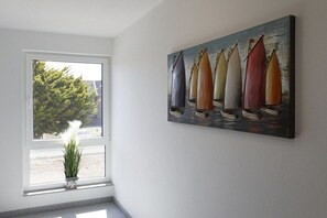 Une pièce avec une grande fenêtre, un tableau représentant des voiliers accroché au mur et une plante en pot posée sur le rebord de la fenêtre.
