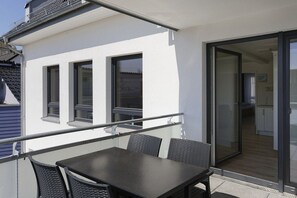 Een balkon met een tafel en stoelen, een schuifdeur naar een keuken, en een witte gevel.
