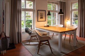 A dining area with a wooden table, white chairs, and a view through large windows. - Holiday home with 1 bedroom (Krummhörn)