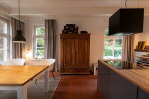 A modern kitchen with a wooden table, white chairs, a cabinet, and a black hanging light fixture. - Holiday home with 1 bedroom (Krummhörn)