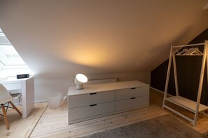 A room with a sloped ceiling, a white dresser with drawers, a small desk with a lamp, and a white coat rack. - Holiday home with 1 bedroom (Krummhörn)
