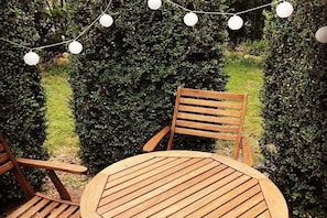 A wooden outdoor seating area with a round table and chairs, surrounded by neatly trimmed hedges and string lights. - Holiday apartment with 3 bedrooms (Neustadt)
