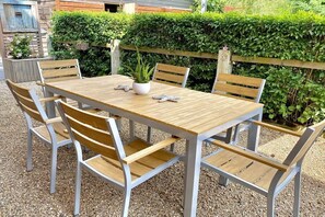 Une table en bois placée à l’extérieur, entourée de six chaises, avec un sol recouvert de gravier, et un arbuste ainsi qu’une clôture en bois en arrière-plan.