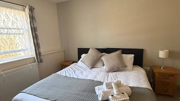 Una cama bien hecha con almohadas y una colcha, una mesita de noche con lámpara, y una ventana con cortinas.
