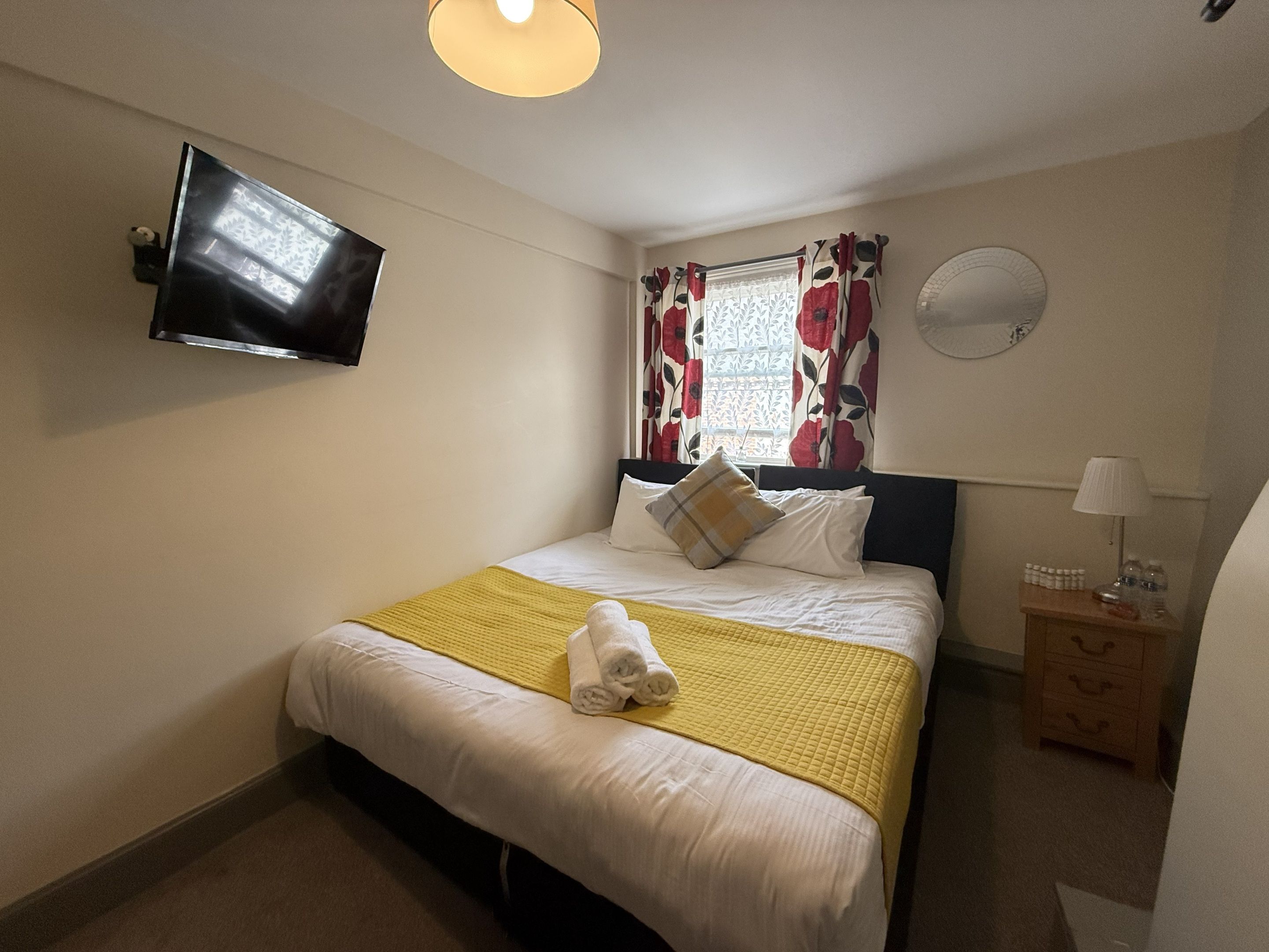 Una camera d'albergo con un letto, una TV a schermo piatto montata a parete, un comodino con una lampada e una finestra con tende a fiori.