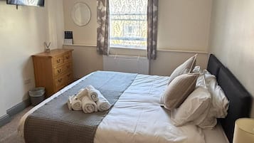 A bedroom with a bed, a wooden dresser, a bedside table, a mirror, and a window with curtains.