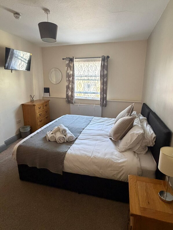A bedroom with a bed, a wooden dresser, a bedside table, a mirror, and a window with curtains.