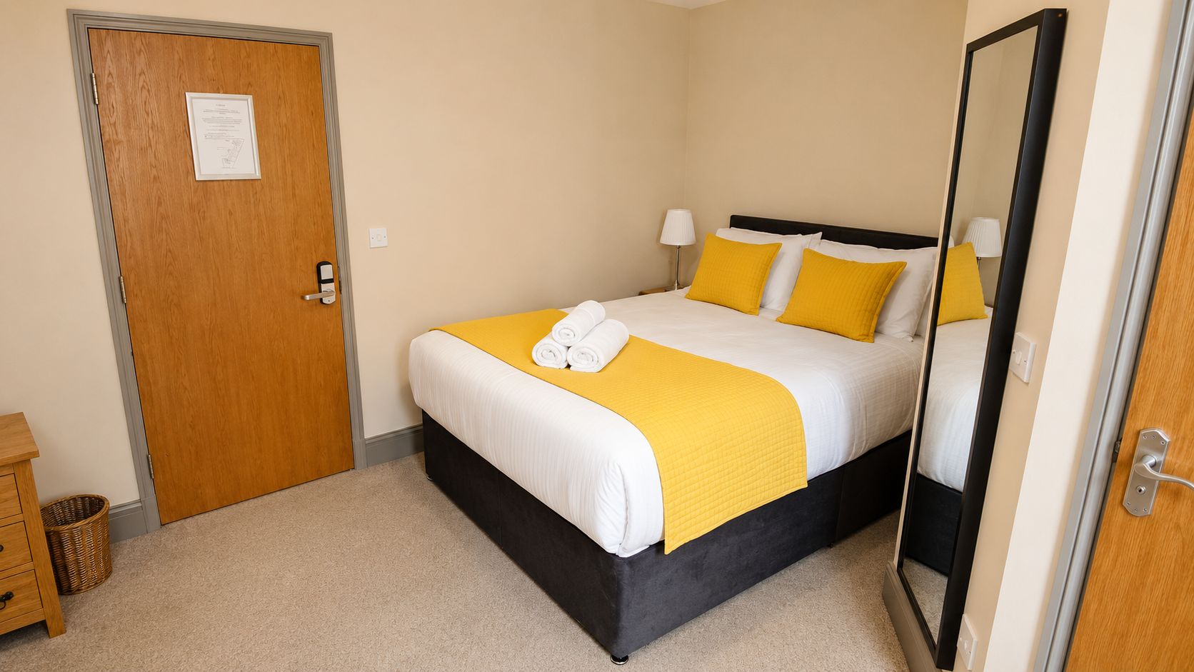A hotel room with a bed, a wooden door, a small bedside table, a lamp, and a mirror.
