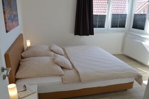 A bed with a headboard, pillows, and a striped blanket. There is a bedside table with a lamp and a book. - New building with dune views (Juist)