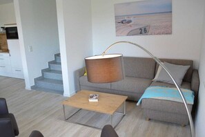 A living room with a sofa, a coffee table, a lamp, a painting, and a staircase leading to a kitchen. - New building with dune views (Juist)