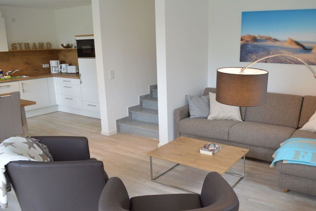 A modern living room with a sofa, armchair, and coffee table. There is a kitchen area with a counter and appliances in the background.