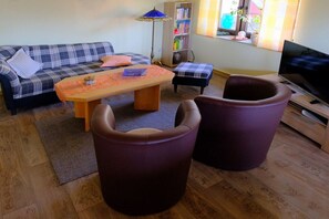 A living room with a sofa, two armchairs, a coffee table, a bookshelf, a TV, and a window with a plant. - Holiday apartment with 1 bedroom (Pottum)