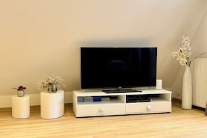 A modern living room with a flat-screen TV on a white stand, a small white side table with a vase, and a tall white vase with flowers. - Holiday apartment in Hambühren with barbecue (Hambühren)