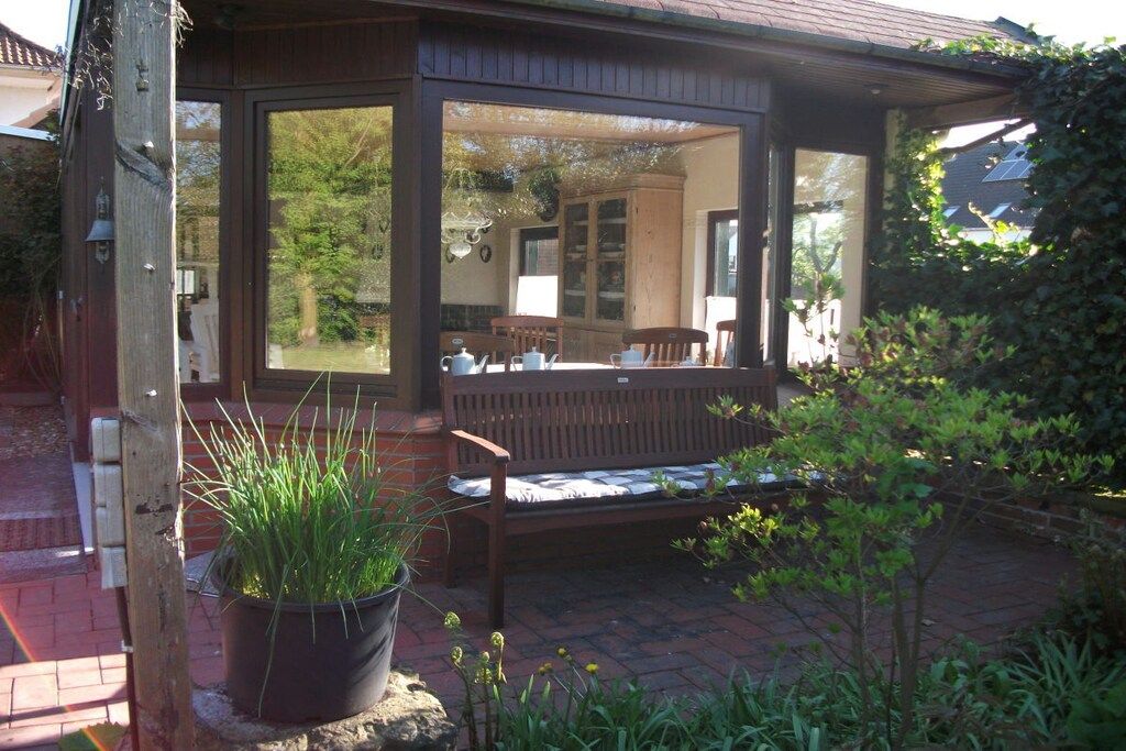 A covered outdoor seating area with a bench, a potted plant, and a view of a house through a glass door.