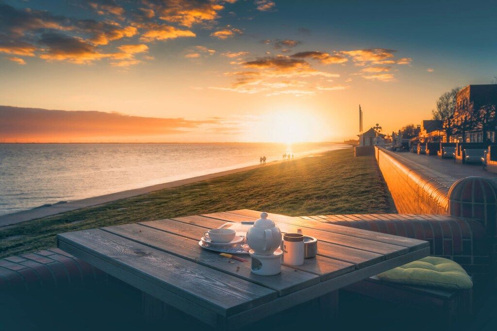A wooden table with a teapot and cups on a beach overlooking the sea at sunset.