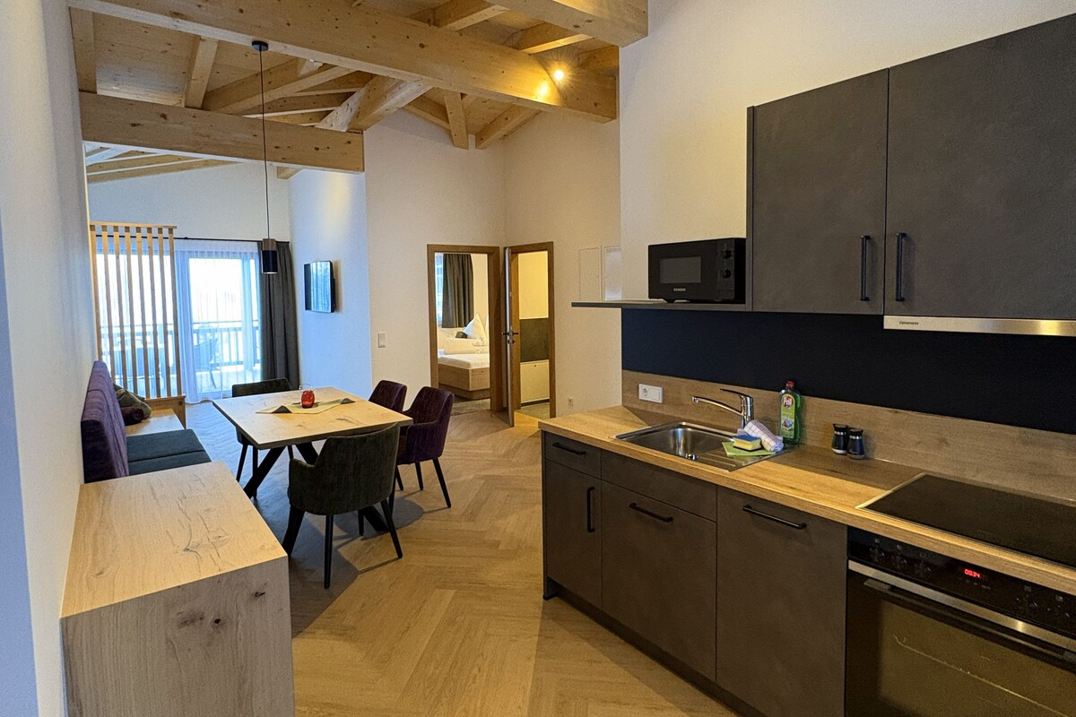 Cozinha moderna com vigas de madeira, área de refeições com mesa e cadeiras, e vista para um quarto com cama e cortinas.