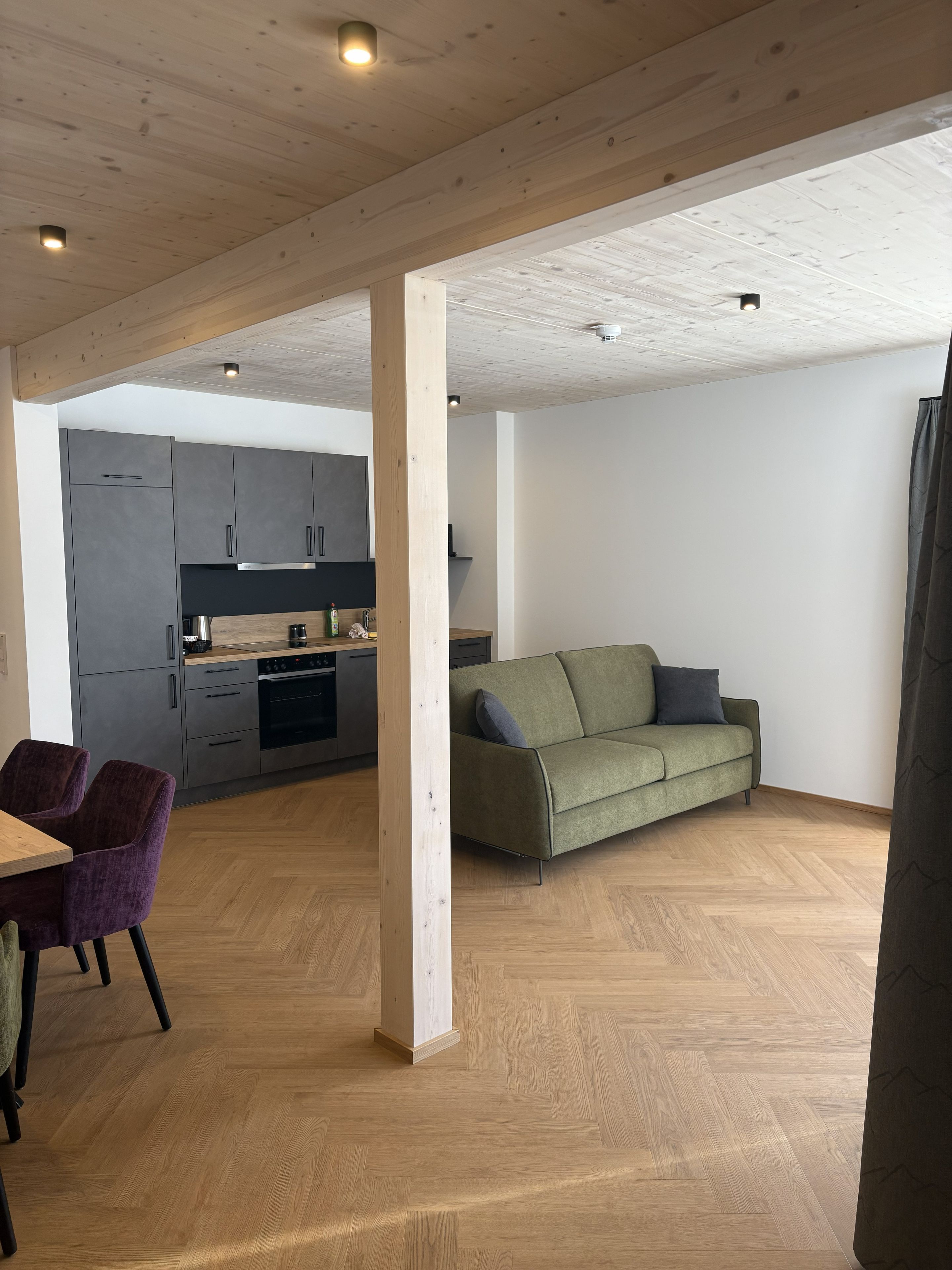 A modern interior with a wooden ceiling, grey kitchen cabinets, a green sofa, and a dining area with purple chairs.