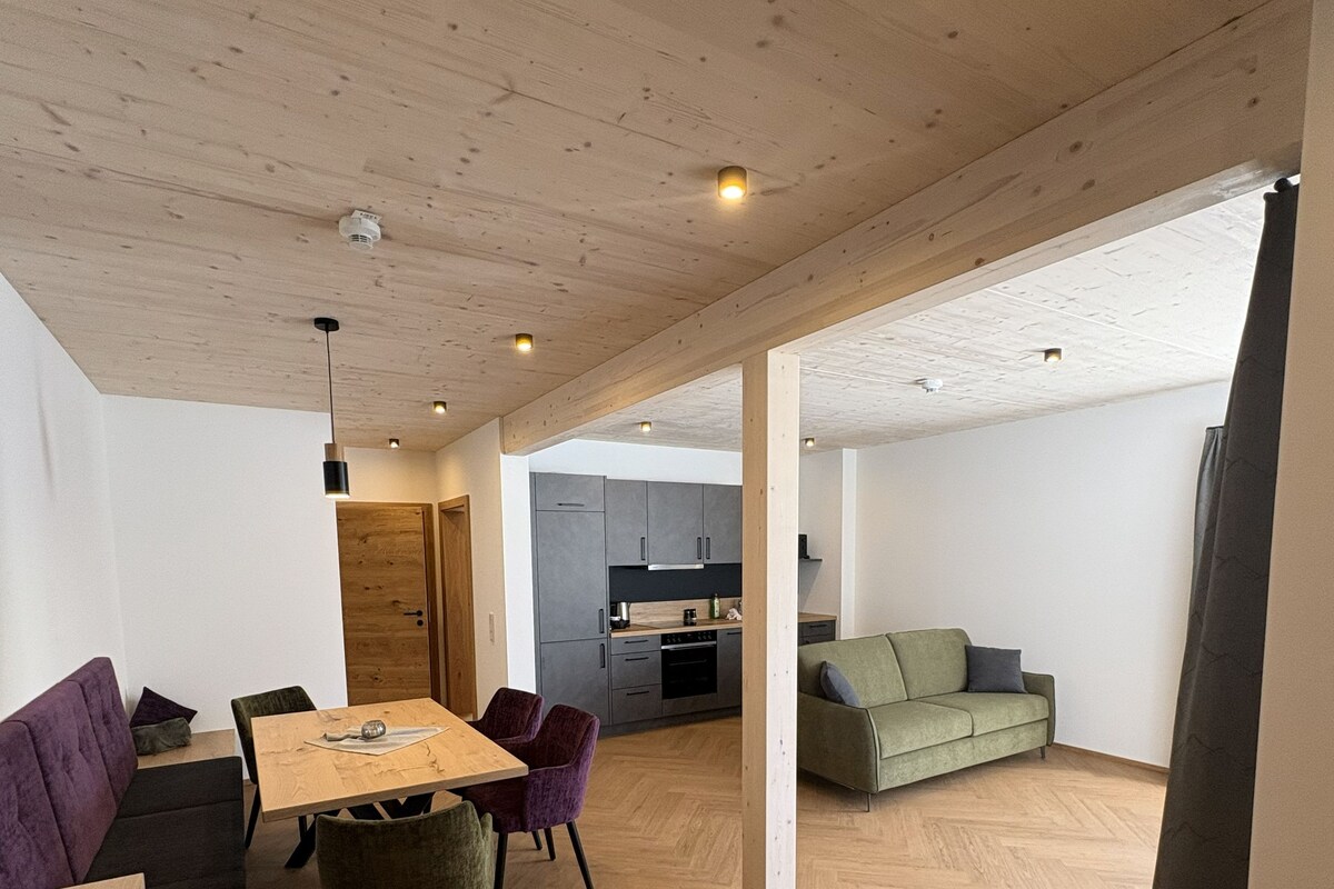 Ampla sala de estar com piso de madeira, mesa de jantar com cadeiras, sofá e cozinha ao fundo.