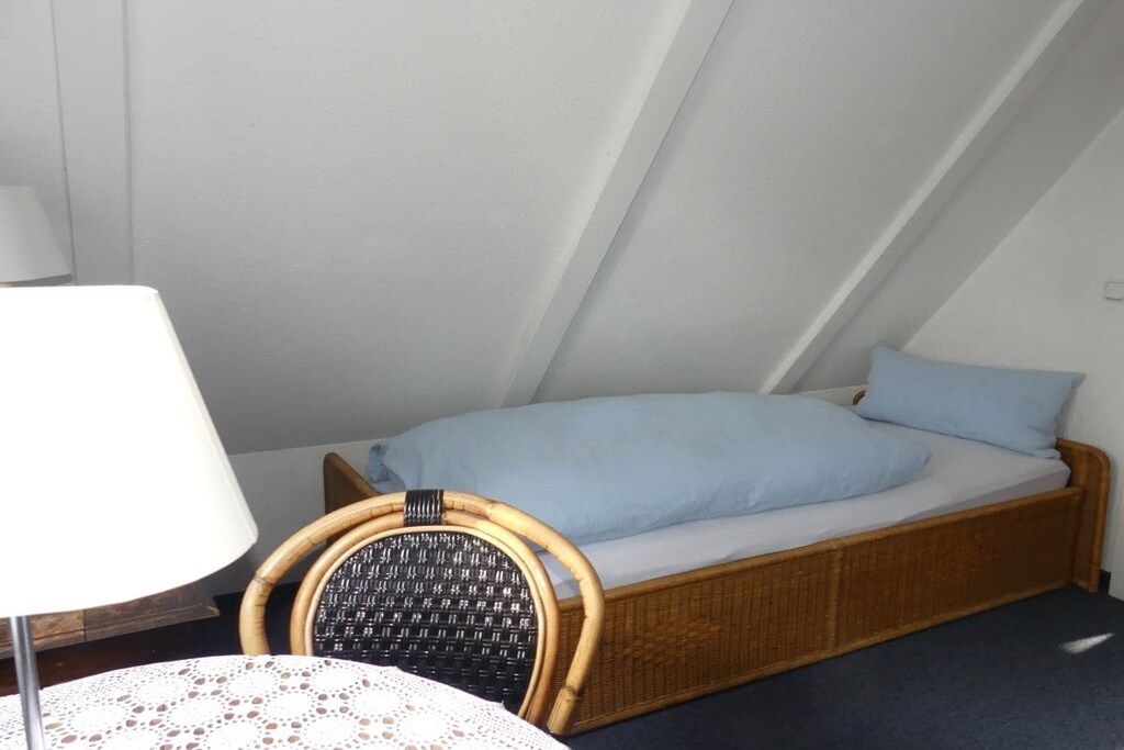 Una habitación pequeña con cama individual, techo inclinado, una silla de mimbre y una mesa con mantel de encaje.